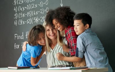 Female teacher smiling and hugging a diverse group of elementary school students in front of a chalkboard