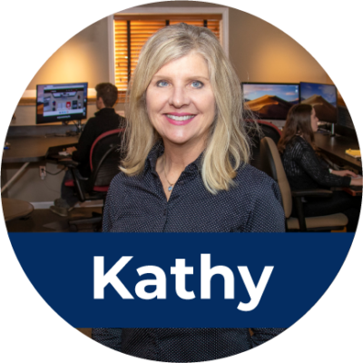 A middle aged white woman with blonde hair stands in an office, smiling. Across the bottom is white text in a blue rectangle that reads "Kathy".