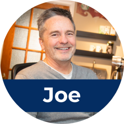 A middle aged white male sits in a a chair and smiles. Across the bottom is white text in a blue rectangle that reads "Joe".