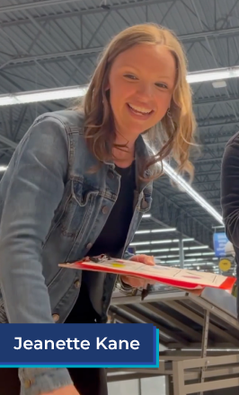 A white woman with medium length red hair stands in a grocery store. She wears a black shirt and jean jacket, and holds a red clipboard. She smiles and is talking. In the bottom left corner is a blue box with white text in it that reads "Jeanette Kane".