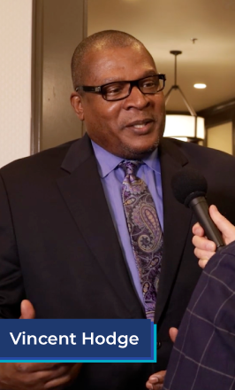 A middle aged African American man stands in the doorway of a conference hall. He wears glasses, a black suit coat, a purple shirt, and a purple paisley tie. In the bottom left corner is a blue box with white text in it that reads "Vincent Hodge".