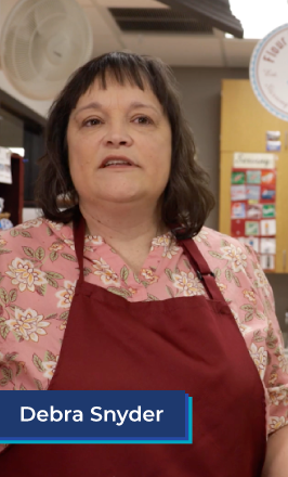 A middle aged white woman with short brown hair stands in a classroom. She wears a pink floral button down shirt underneath a red apron. In the bottom left corner is a blue box with white text in it that reads "Debra Snyder".