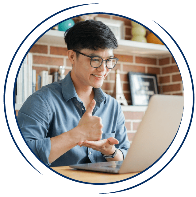 In a circular frame: A young Asian man sits at a table in front of a laptop. He is communicating with sign language and smiling at the screen of the computer.