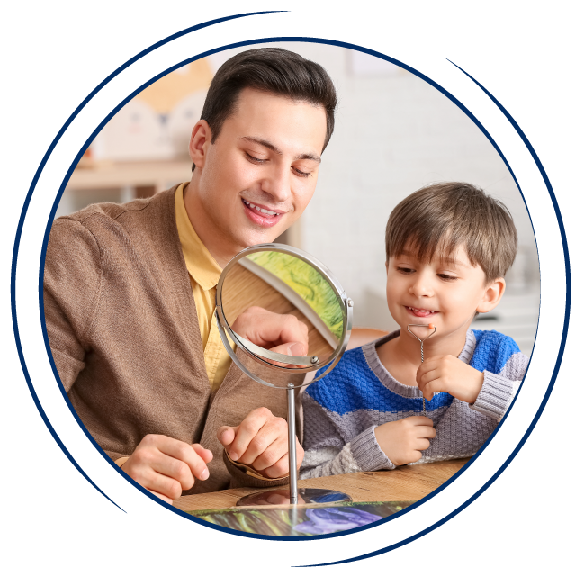 In a circular frame: A young white man and a young white boy sit at a wooden table in a classroom. They look into a small mirror together. They boy is holding a sensory toy, which is resting on his chin.