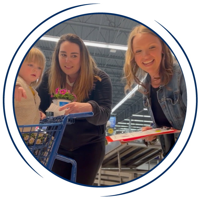 In a circular frame: Two white adult women and a young white girl stand in a grocery store looking at fruit. The girl sits in a shopping cart.