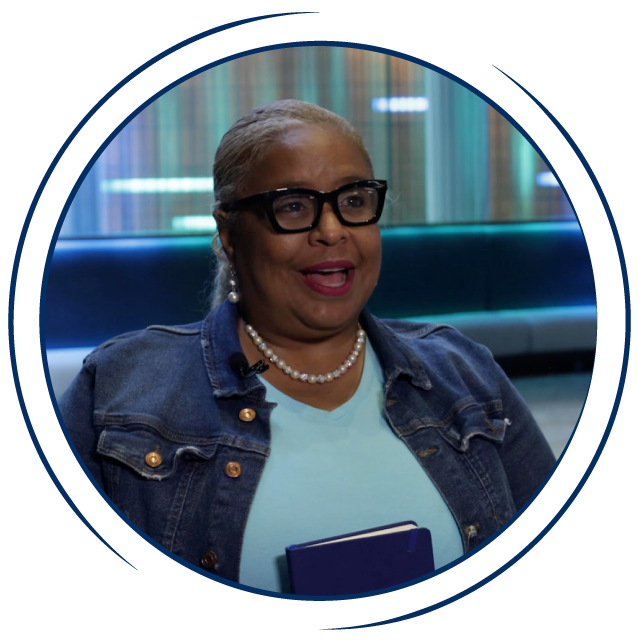In a circular frame:A middle aged African American woman sits in the lobby of a hotel. She wears black glasses, a blue tee shirt, a jean jacket, and a pearl necklace. She holds a blue planner, and smiles while speaking.