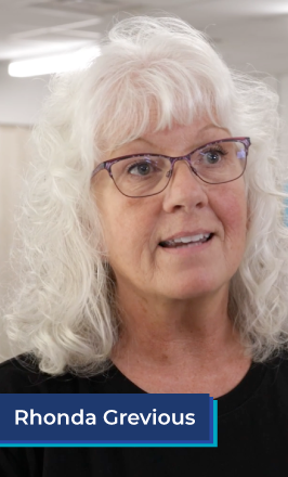 An older white woman with long white hair and pink glasses stands in a classroom. She wears a black tee shirt, and smiles while talking. In the bottom left corner is a blue box with white text in it that reads "Rhonda Grevious".