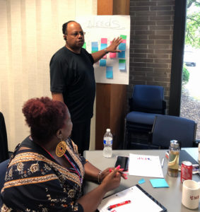 A middle aged African American man stands in front of a conference table, where an African American woman with red hair sits. The man stands near an idea board with colorful post-it notes on it. He is speaking and gesturing to the board.
