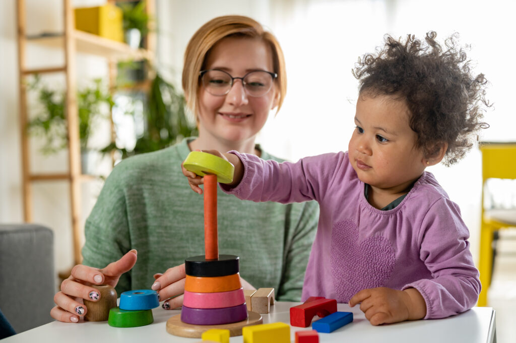 A young child plays with colorful stacking rings while an early intervention specialist watches and assists.