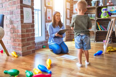 A school social worker sitting on the floor with a clipboard, engaging with a young child in a classroom filled with toys and colorful drawings.