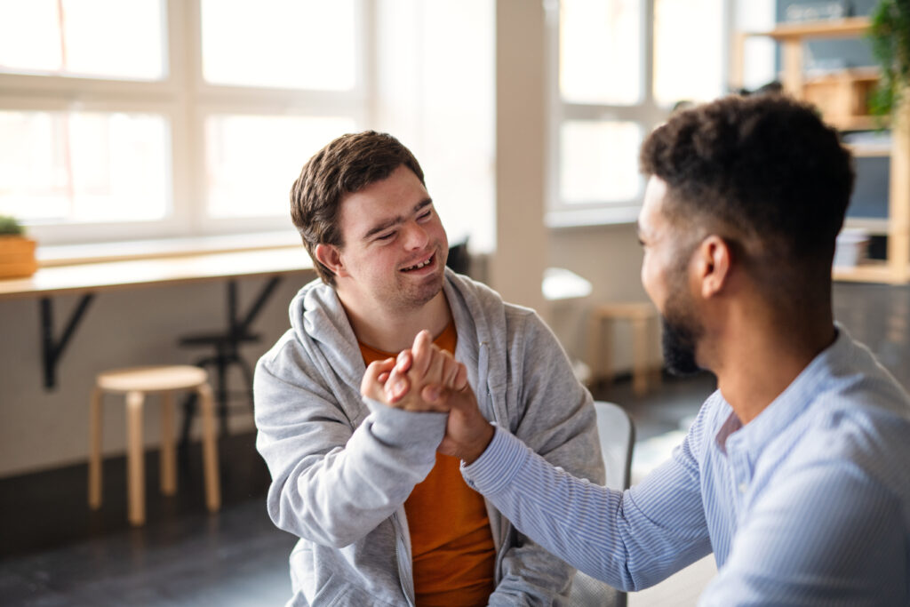 Young man with a disability smiling and engaging in a handshake with a support worker in a bright room.