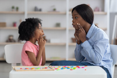 Speech-language pathologist having lesson with little girl, working on pronunciation, touching faces and grimacing.