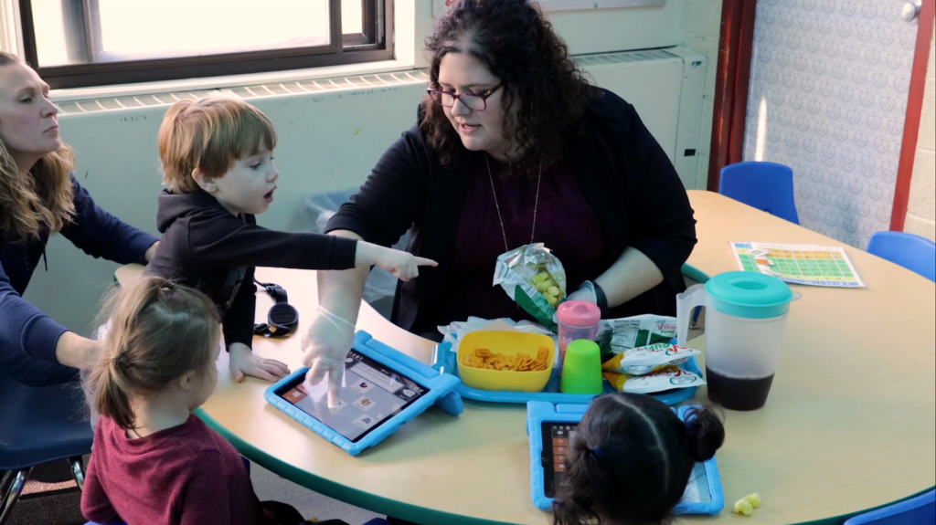 Teacher helping young children with disabilities use assistive technology on tablets during a classroom activity.